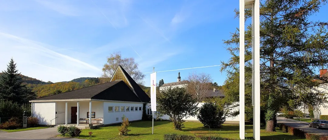 Eine moderne weiße Kirche mit einem Glockenturm und einem Kreuz oben, vor einem blauen Himmel mit Wolken, umgeben von Grün.