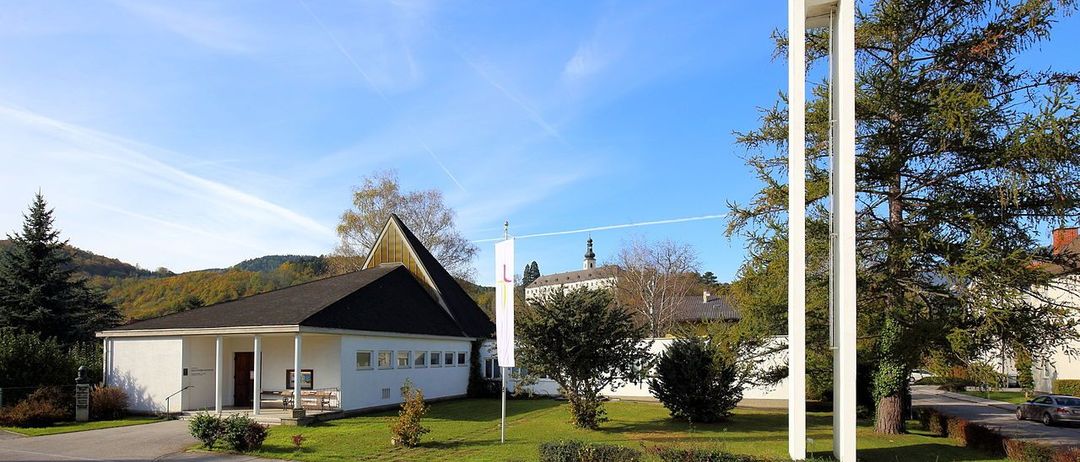 Eine moderne weiße Kirche mit einem Glockenturm und einem Kreuz oben, vor einem blauen Himmel mit Wolken, umgeben von Grün.