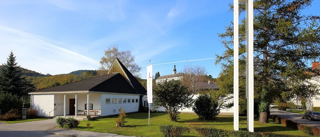 Eine moderne weiße Kirche mit einem Turm und einem Kreuz, vor einem klaren blauen Himmel und umgeben von Grün.