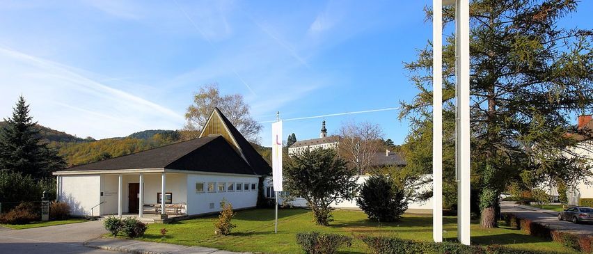 Eine moderne weiße Kirche mit einem Turm und einem Kreuz, vor einem klaren blauen Himmel und umgeben von Grün.