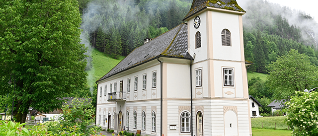Eine traditionelle Kirche mit einem Turm und einer Uhr, umgeben von üppigem Grün und Bergen im Hintergrund.