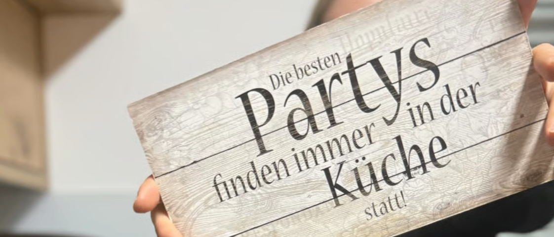 Ein Holzschild mit dem Text 'Partys finden immer in der Kuche statt!' wird von einer Person in einem Raum gehalten.