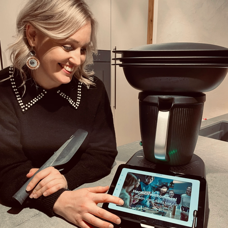 Eine Frau mit einem Messer zeigt auf ein Tablet mit einem Rezept. Sie lächelt und scheint in einer Küche zu sein.