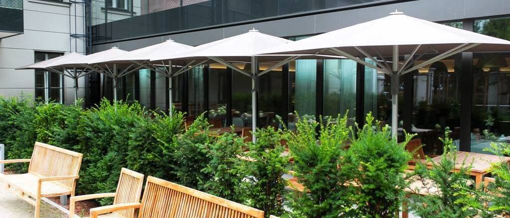 Terrasse eines Restaurants mit Holzbänken und großen weißen Sonnenschirmen, umgeben von Hecken und einem modernen Gebäude im Hintergrund.