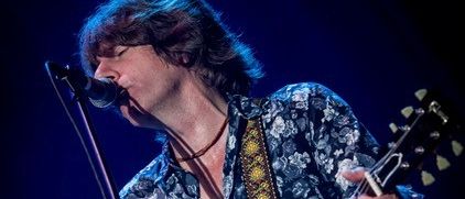 Ein Mann mit langen Haaren und einem Blumenhemd spielt eine Gitarre und singt in ein Mikrofon auf der Bühne.