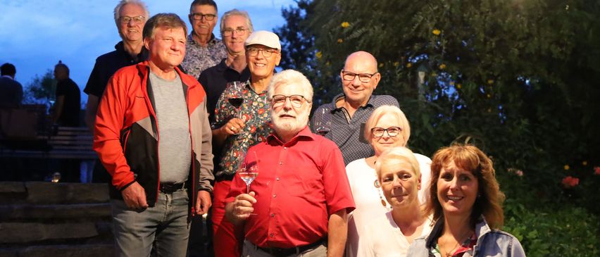 Eine Gruppe lächelnder Erwachsener, einige mit Weinglas in der Hand, posiert für ein Foto im Freien vor einem Baumhintergrund.