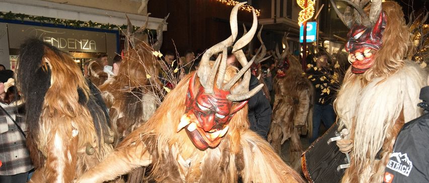 Eine Gruppe von Menschen in Pelzkostümen mit Hörnern und Masken tanzt bei Nacht auf der Straße. Im Hintergrund sind Gebäude und Weihnachtsbeleuchtung zu sehen.