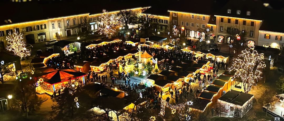 Ein Nachtblick aus der Luft auf einen festlich geschmückten Stadtplatz, der mit funkelnden Weihnachtslichtern und einer Vielzahl von Ständen und Zelten verziert ist. Die Menschen versammeln sich, um die weihnachtliche Atmosphäre zu genießen.