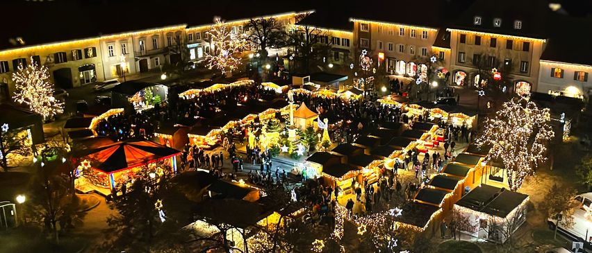 Ein Luftbild eines geschäftigen Weihnachtsmarktes bei Nacht mit vielen Ständen, festlichen Lichtern und Menschen, die herumlaufen. Die Gebäude rundherum sind ebenfalls beleuchtet, und im Hintergrund befindet sich eine Kirche.