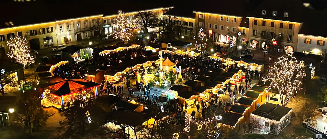 Ein nächtlicher Luftblick auf einen geschäftigen Weihnachtsmarkt, gefüllt mit festlichen Lichtern und Dekorationen, auf einem europäischen Stadtplatz.