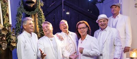 Eine Gruppe von Männern in weißen Anzügen posiert für ein Foto vor einer Statue. Sie lächeln und scheinen Spaß zu haben. Der Mann in der Mitte hält eine Halskette.