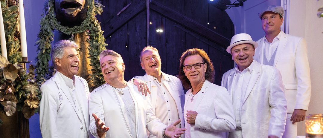 Eine Gruppe von Männern in weißen Anzügen posiert für ein Foto vor einer Statue. Sie lächeln und scheinen Spaß zu haben. Der Mann in der Mitte hält eine Halskette.