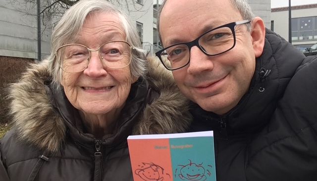 Eine ältere Frau und ein Mann lächeln und halten ein Buch mit dem Titel 'Wie Kannst Du Nur...'. Sie tragen warme Jacken und stehen vor einem Gebäude.