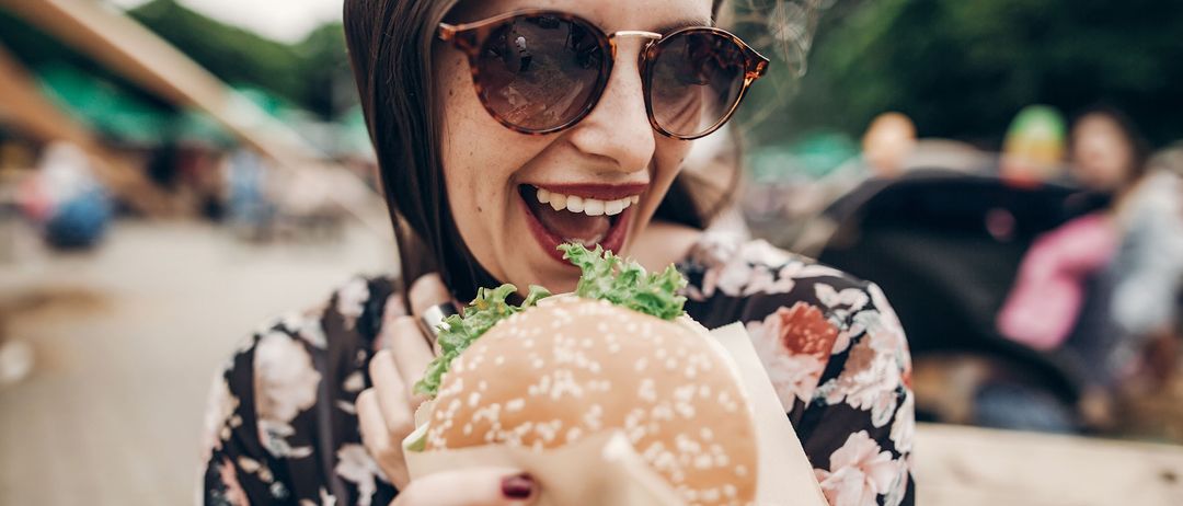 A woman in sunglasses is holding a hamburger with lettuce and smiling while eating it. She is wearing a floral shirt and appears to be enjoying her meal.