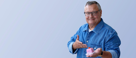 Ein Mann in einem Jeanshemd gibt ein Daumen hoch und hält eine rosa Süßigkeit. Er trägt eine Brille und einen Ring.