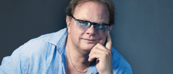 Ein Mann sitzt auf einem Stuhl, hält eine Gitarre in der Hand, trägt ein blaues Hemd, eine Halskette und eine Brille. Der Text 'Live is Life' ist auf dem Bild zu sehen.