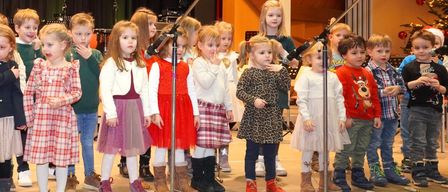 Mehrere kleine Kinder stehen auf einer Bühne, gekleidet in verschiedene Winteroutfits, möglicherweise bei einer Gesangsaufführung. Ein Mikrofon auf einem Ständer steht vor ihnen.