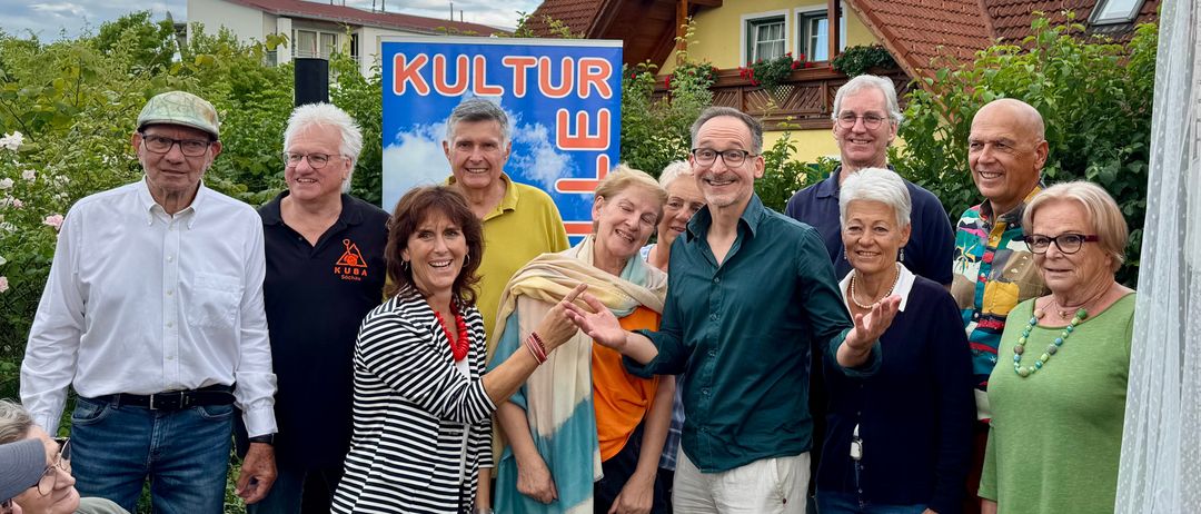Eine Gruppe von Menschen steht draußen, lächelt und posiert für ein Foto vor einem Haus mit einem Schild, das 'Kultur' sagt.