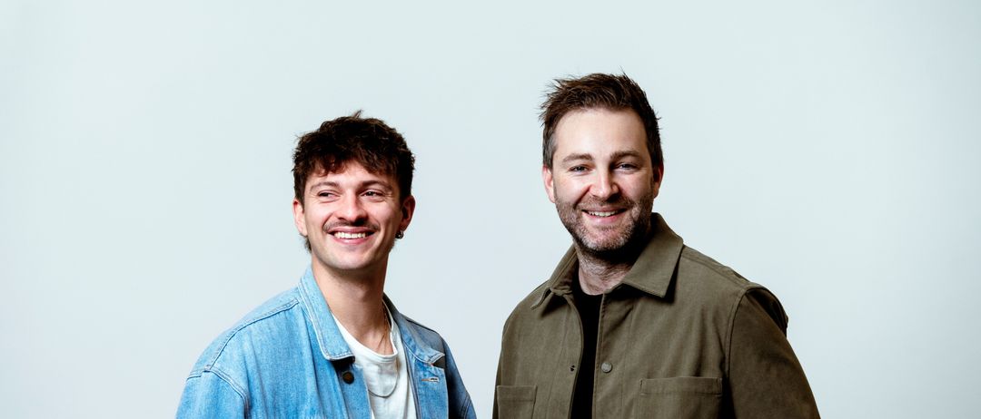 Two men standing next to each other, smiling for a photo. The man on the left wears a denim jacket and a white shirt. The man on the right wears a green jacket with a beard.