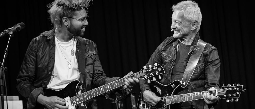 Zwei Männer spielen Gitarren auf einer Bühne. Der linke Mann hat eine unordentliche Frisur und trägt eine Halskette, während der rechte Mann lächelt und eine Lederjacke trägt.