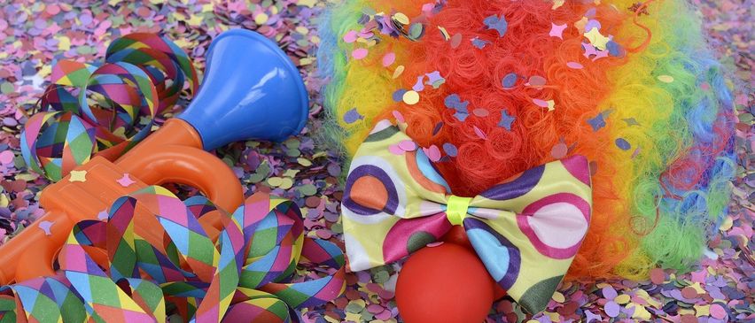 Eine Clownperücke, ein roter Ball und eine blaue Hupe liegen auf einem Bett aus bunten Konfetti und Luftschlangen.
