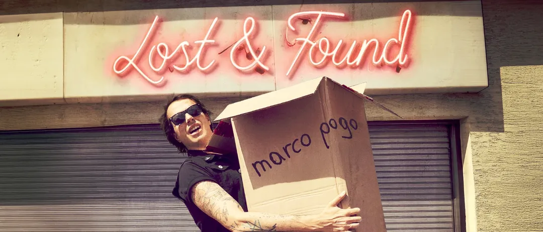 Ein Mann mit Tätowierungen an den Armen trägt einen Karton mit der Aufschrift 'marco pogo'. Er steht vor einem Gebäude mit einem Neonschild, das 'Lost & Found' besagt.