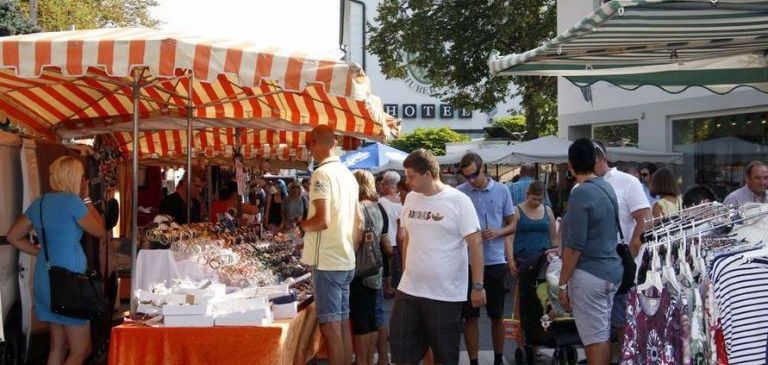 At a market with awnings, people shop for various items. A woman stands by a table selling jewelry, while others browse through clothing on hangers. A building with a sign reading Hotel is visible in the background.