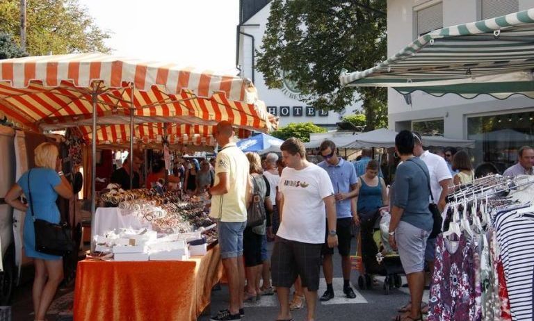 Auf einem Markt mit Markisen kaufen Menschen verschiedene Artikel. Eine Frau steht an einem Tisch, der Schmuck verkauft, während andere Kleidung an Kleiderbügeln durchstöbern. Im Hintergrund ist ein Gebäude mit einem Schild zu sehen, das Hotel steht.