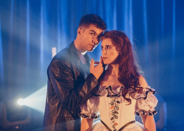 Ein Mann und eine Frau stehen eng beieinander auf der Bühne und tragen formelle Kleidung. Der Mann trägt eine schwarze Lederjacke, während die Frau ein weißes Kleid mit Blumenmuster trägt. Sie schauen sich beide an und posieren möglicherweise für ein Foto.