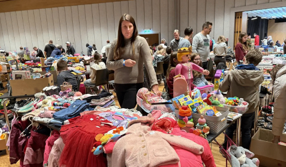 Eine Frau steht in einem großen Innenbereich, umgeben von Tischen, die mit Spielzeug, Kleidung und anderen Gegenständen gefüllt sind. Menschen durchstöbern die Artikel, und einige stehen und unterhalten sich. Der Raum hat einen Holzboden und weiße Wände.