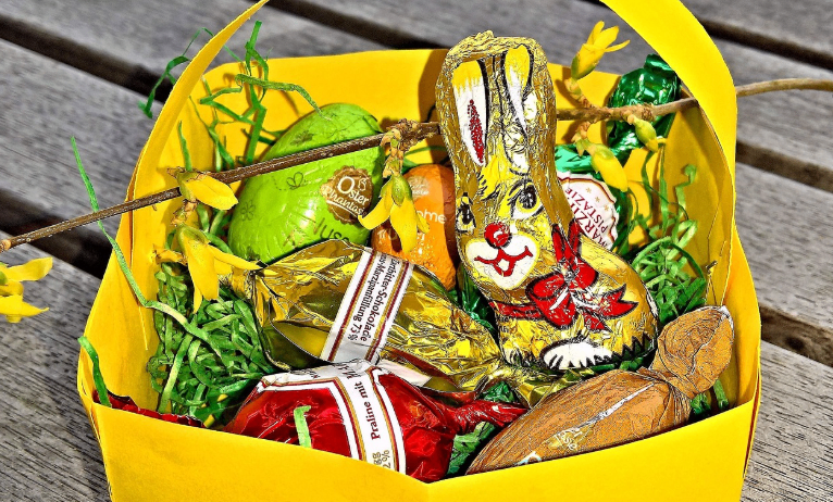 Ein gelber Osterkorb gefüllt mit verschiedenen Osterschokoladen, darunter ein Hase, in Goldfolie gewickelt, auf einer Holzoberfläche platziert.