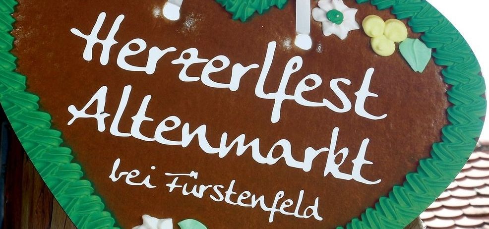 Ein herzförmiger Lebkuchen mit grünen Blättern und weißen Blumen drumherum sagt 'Herzfest Altenmarkt bei Fürstenfeld' in weißen Buchstaben.