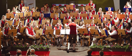 A large orchestra is performing on a stage with various musical instruments, accompanied by a conductor, under a red carpet and surrounded by seated audience members.