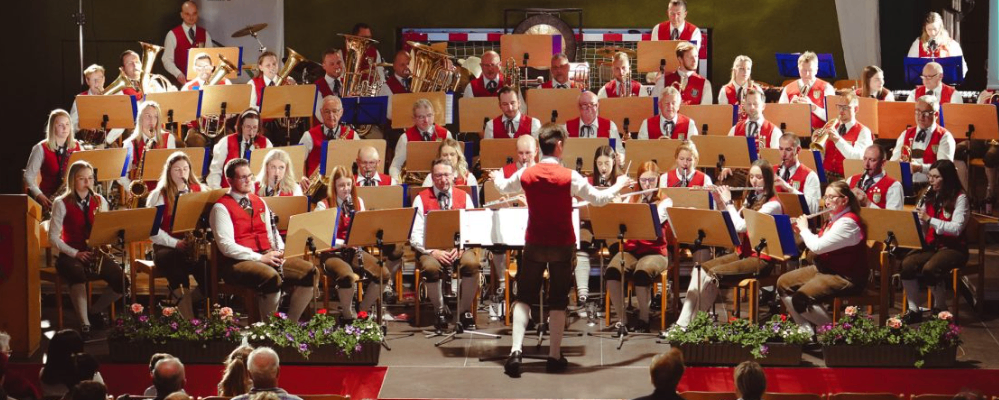 Ein großes Orchester spielt auf der Bühne, mit Musikern, die verschiedene Instrumente spielen, einschließlich Flöten und Posaunen, während ein Dirigent führt. Das Publikum sitzt vor ihnen.