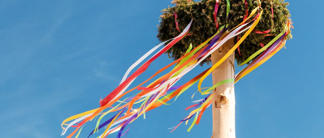 Ein hoher Holzpfahl mit einem Kranz aus immergrünen Pflanzen oben und bunten Bändern, die davon herabhängen, vor einem klaren blauen Himmel.