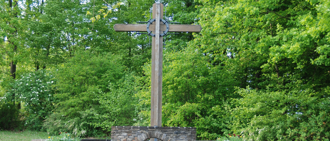 Ein großes hölzerne Kreuz steht an einem Gedenkort, umgeben von üppigem Grün und Stufen, die zu ihm hinaufführen. Zwei Steinblöcke auf beiden Seiten haben Tafeln mit Inschriften.