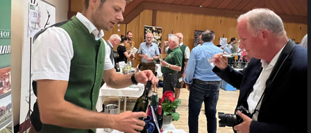 Ein Mann in grüner Weste und brauner Hose schenkt Wein an einem Tisch mit einem Eimer Weinflaschen ein. Ein anderer Mann probiert den Wein und hat eine Kamera um die Schulter.