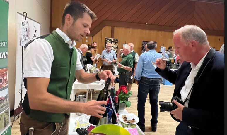 Ein Mann in grüner Weste und brauner Hose schenkt Wein an einem Tisch mit einem Eimer Weinflaschen ein. Ein anderer Mann probiert den Wein und hat eine Kamera um die Schulter.