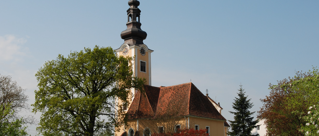 Eine traditionelle Kirche mit braunem Dach und einem Turm mit Uhr steht zwischen Bäumen und Gras unter einem klaren blauen Himmel.