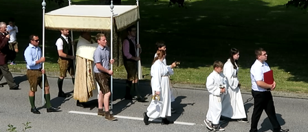 Eine Gruppe von Menschen in traditioneller bayerischer Kleidung geht mit einem Baldachin auf der Straße, möglicherweise für eine religiöse Prozession.