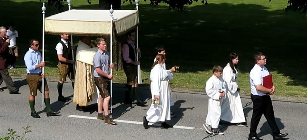 A group of people in traditional Bavarian clothing are walking on the road with a canopy, possibly for a religious procession.