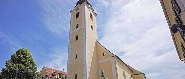 Ein hoher, schmaler Turm steht neben einer Kirche mit rotem Dach. Eine Bank und eine Straßenlampe sind draußen. Bäume und Gras umgeben das Gebiet.