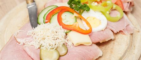 A wooden chopping board displays a variety of ingredients including ham, cheese, cucumber, bell peppers, tomatoes, and a half-boiled egg, accompanied by a knife.