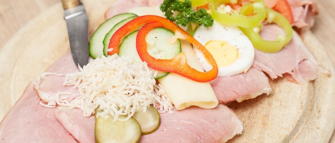 Ein hölzernes Schneidebrett zeigt eine Vielzahl von Zutaten, darunter Schinken, Käse, Gurken, Paprika, Tomaten und ein halb gekochtes Ei, begleitet von einem Messer.