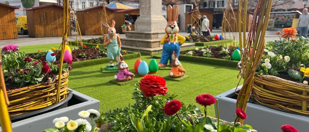 Ein Garten voller Osterdekorationen mit Hasenfiguren, bunten Eiern und Blumen. Ein Denkmal steht in der Mitte, mit Gebäuden und Bäumen im Hintergrund.