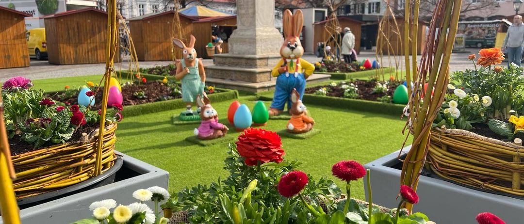 Ein Garten mit künstlichen Hasen, bunten Eiern und Blumen. Im Hintergrund befinden sich Holzstände und Gebäude.