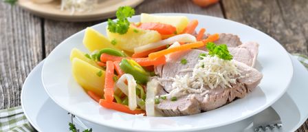 A white plate with a sliced meat dish topped with cheese and garnish, accompanied by a mix of vegetables such as carrots, potatoes, and green peppers. A cheese grater and more vegetables are in the background.