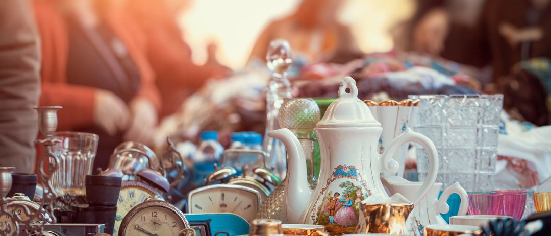 Ein überladener Tisch mit antiken Teekannen, Uhren und verschiedenen Gegenständen, Personen im Hintergrund.