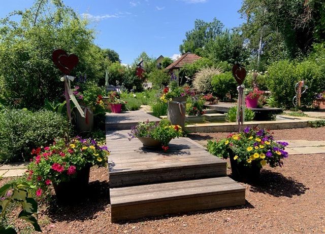 A wooden deck with vibrant flowers and plants, surrounded by greenery and heart-shaped decorations. The garden features a pond, trees, and a house in the background.