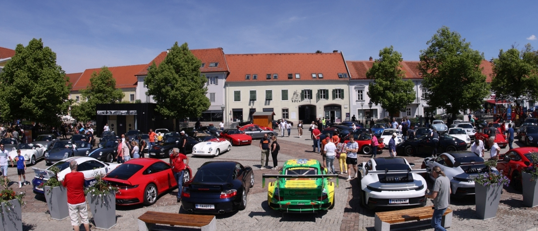 Eine große Ansammlung von Autos ist auf einer gepflasterten Straße geparkt, mit Menschen, die herumlaufen und sie bewundern. Im Hintergrund befindet sich ein Gebäude mit einem roten Dach und großen Fenstern.